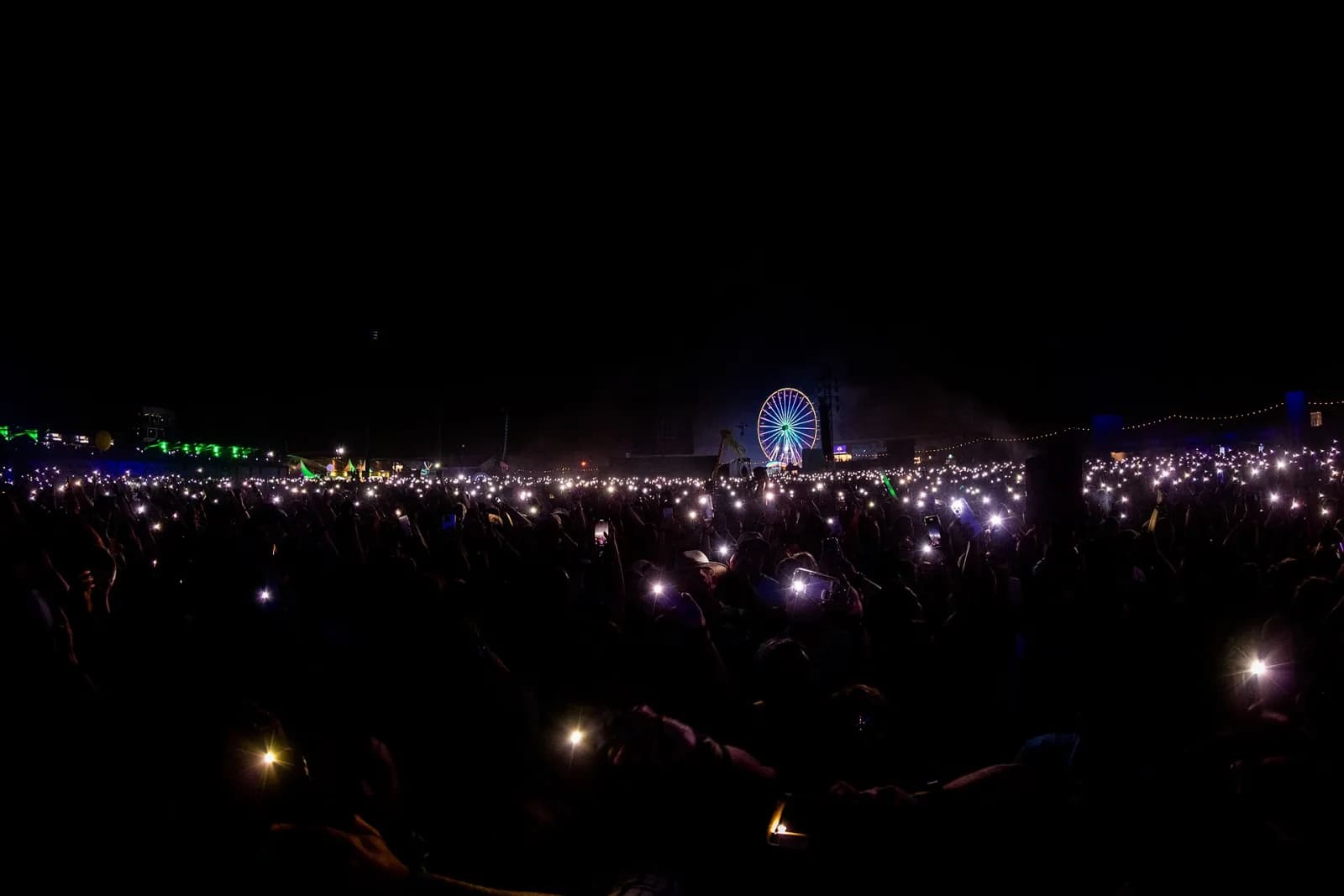 A sea of phone lights in a night-time festival crowd with an illuminated Ferris wheel on the horizon