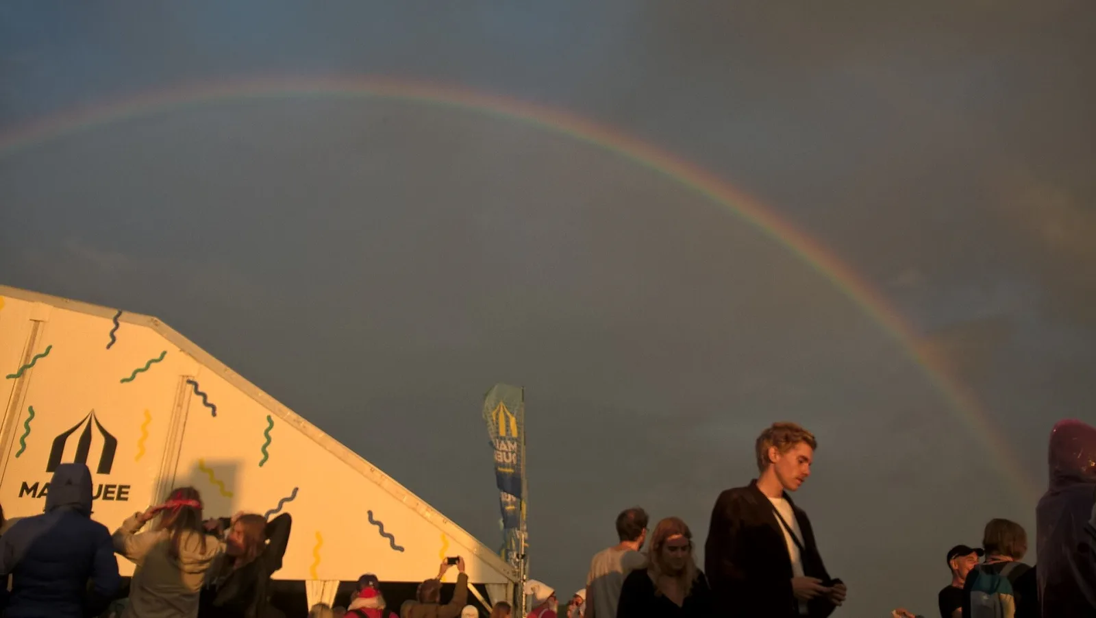 A rainbow arcing over the Pukkelpop site at golden hour, festivalgoers by the Marquee tent