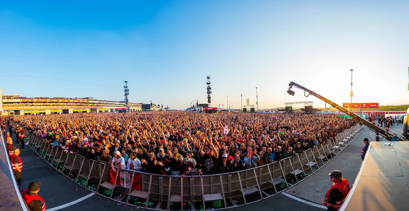 A golden-hour panorama of the Rock am Ring crowd from the front barrier, grandstands behind