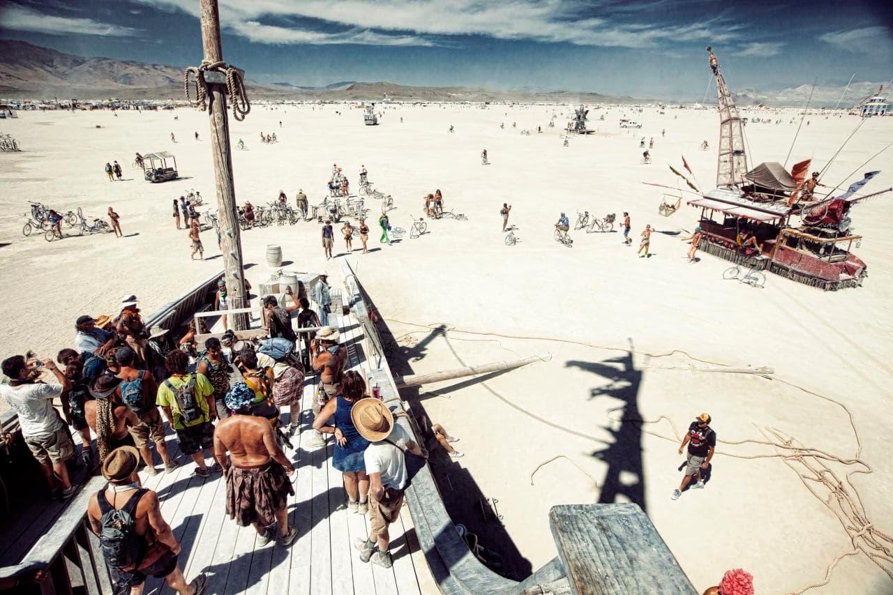 Daytime on the open playa at Burning Man: people on a wooden art structure, cyclists and an art car spread across the dust toward distant mountains