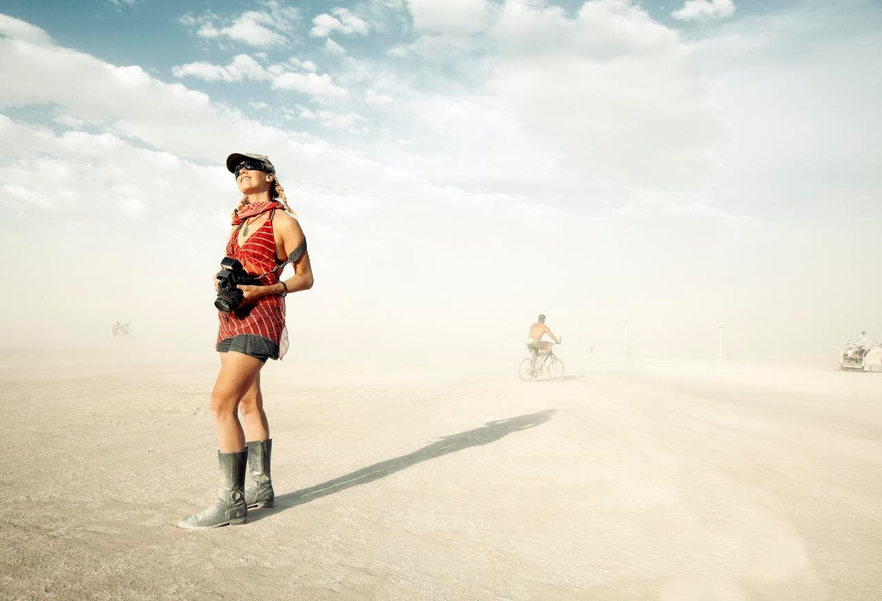 A burner in goggles and a bandana stands on the dusty playa with a camera, a cyclist and an art car hazy in the dust behind her