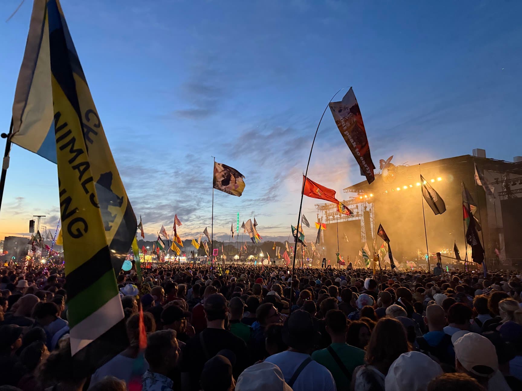 A packed Glastonbury crowd with flags in front of a lit stage at dusk